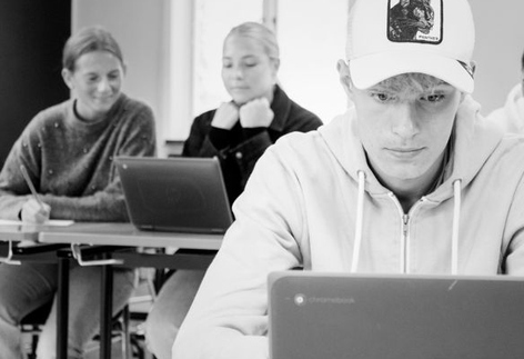Kille i keps sitter framför laptop i ett klassrum. I bakgrunden sitter två elever vid en dator.  elever.  